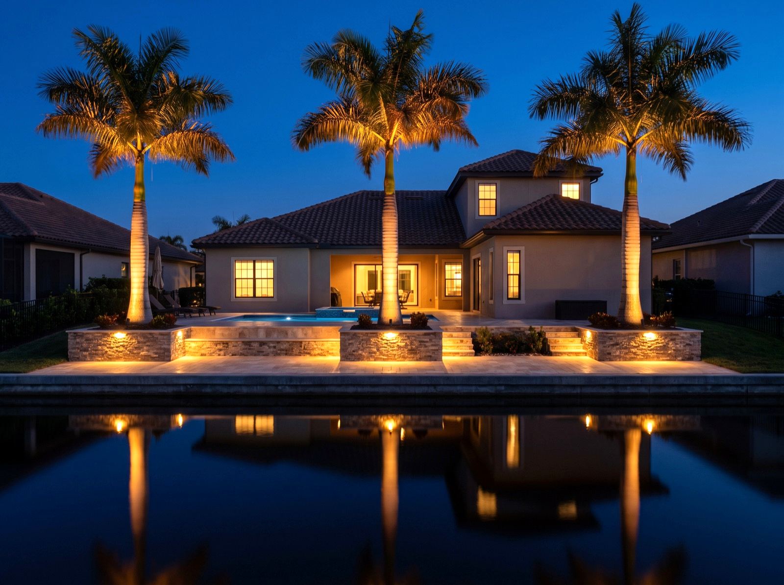 A canal-front property lit at blue hour, seen across still water with palms uplit against a navy sky